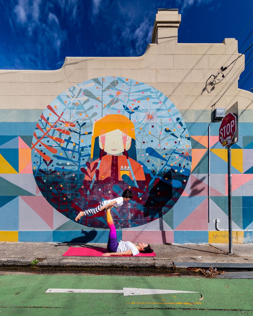 A woman balances in an acrobatic yoga pose on a pink mat in front of a colourful geometric street mural of a figure in an orange hat.