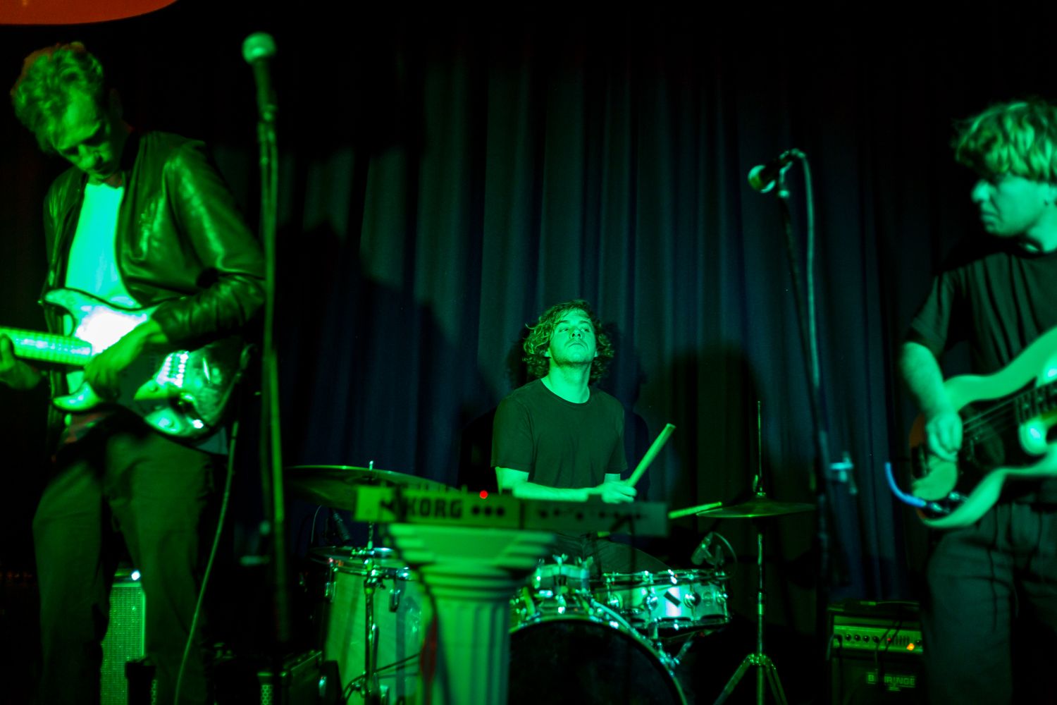 Live band performing on a dimly lit stage with green lighting, featuring a guitarist, a drummer behind a drum kit, and a bassist.