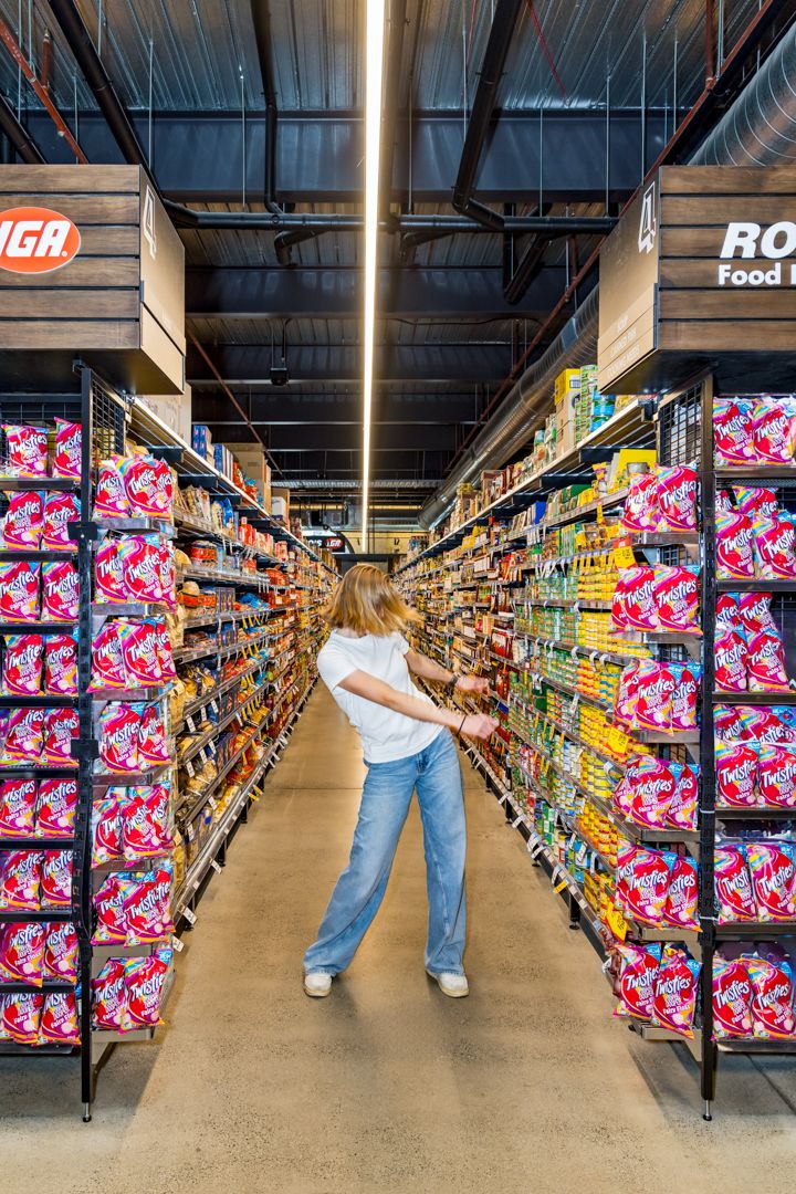 Young woman in jeans stands in a supermarket aisle lined with colorful packaged snacks and groceries.