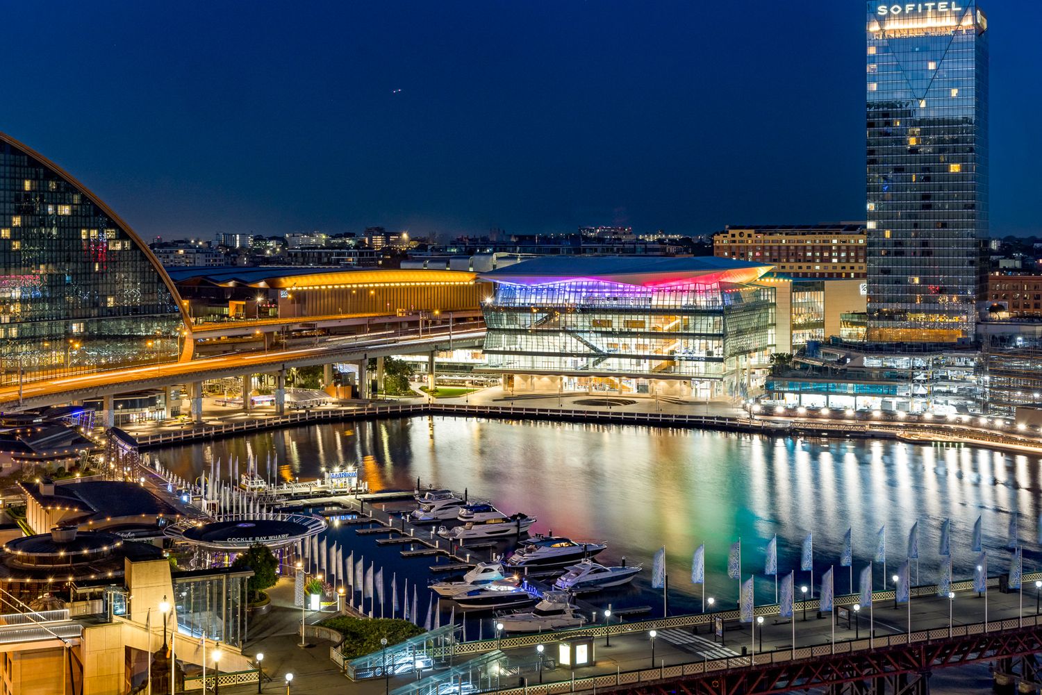 Illuminated waterfront buildings and moored boats reflect on calm harbor water at night beneath a deep blue twilight sky.