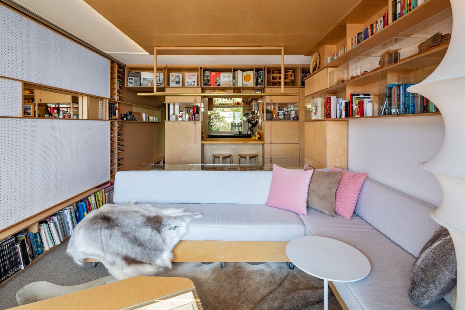 Cozy living room with built-in timber bookshelves, a pale built-in sofa with pink cushions, a sheepskin throw and a round white side table.
