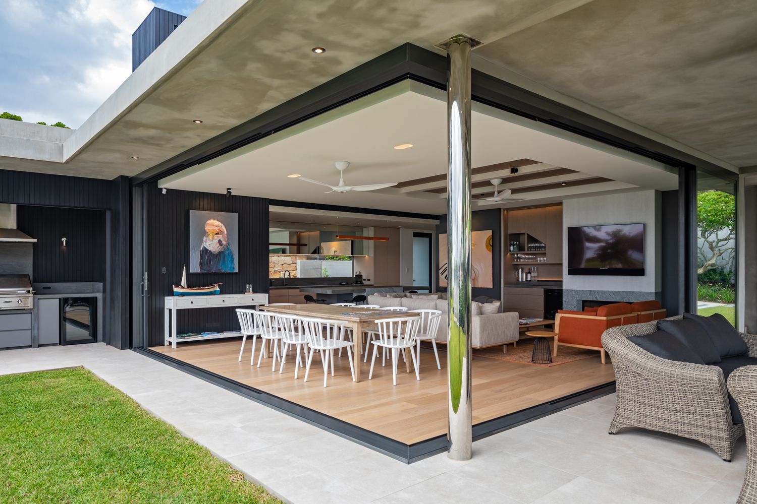 Open-plan indoor-outdoor living area with a covered patio, dining table, lounge seating and sliding glass doors opening to a green lawn.