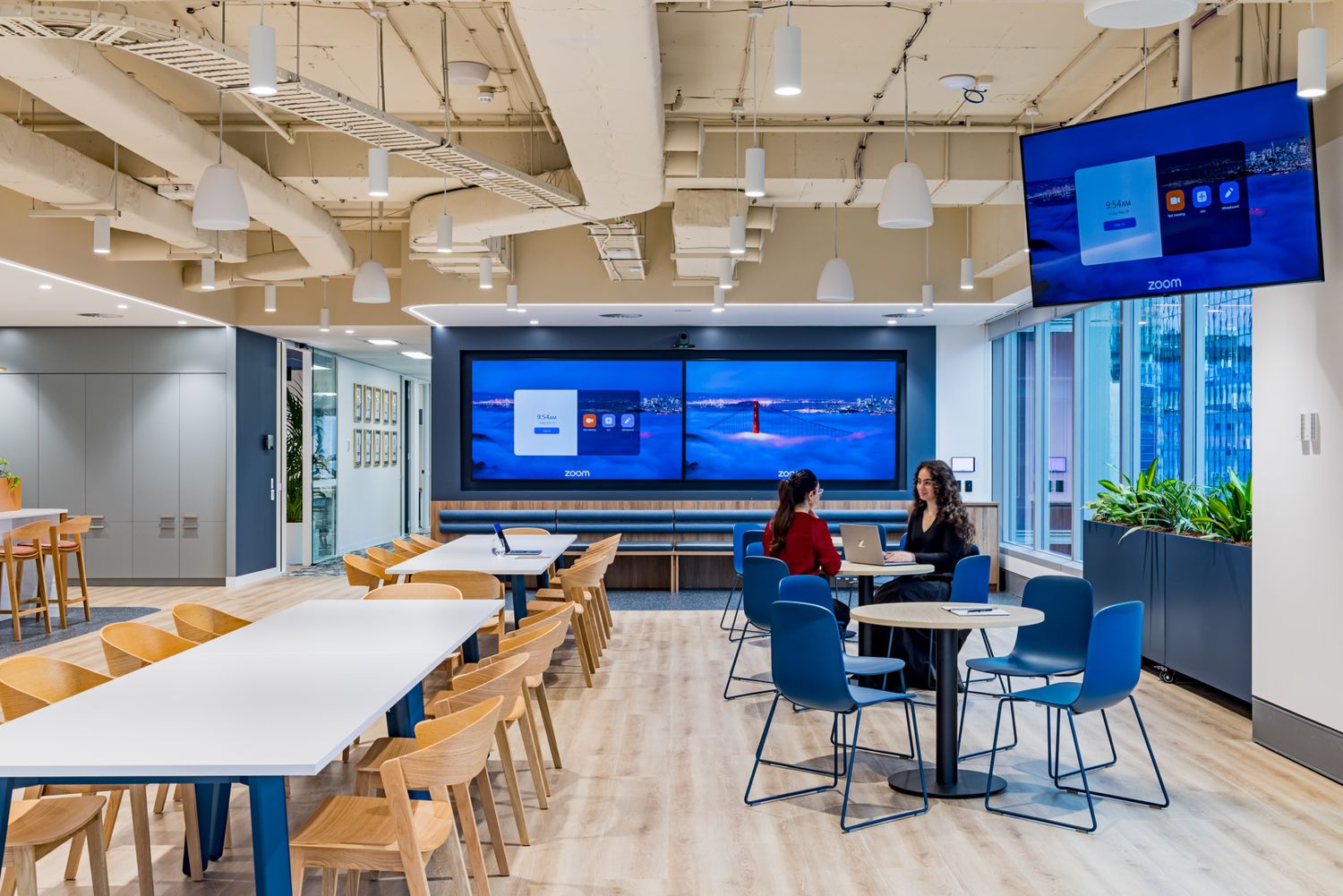 Modern open-plan office breakout area with wooden tables, blue chairs, wall-mounted screens and two colleagues talking.