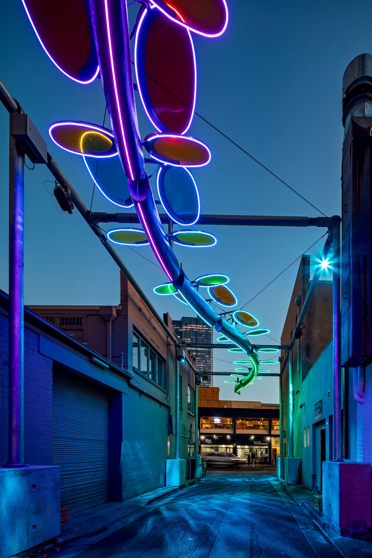 Blue and purple illuminated serpent sculpture with oval discs stretching above a narrow brick alley in the evening.