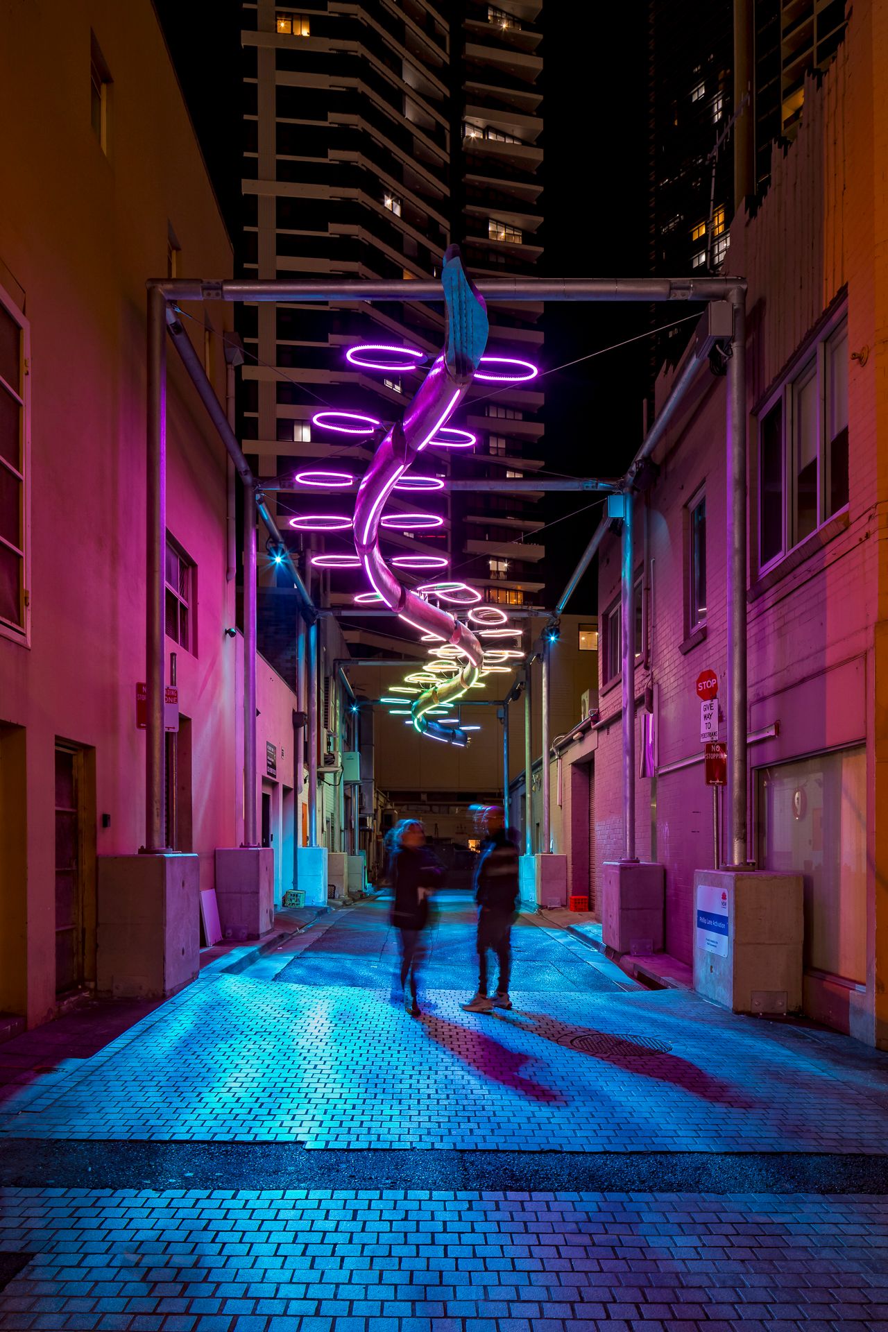 Two pedestrians walk down a narrow city alley at night beneath a glowing purple spiral light installation and tall apartment towers.
