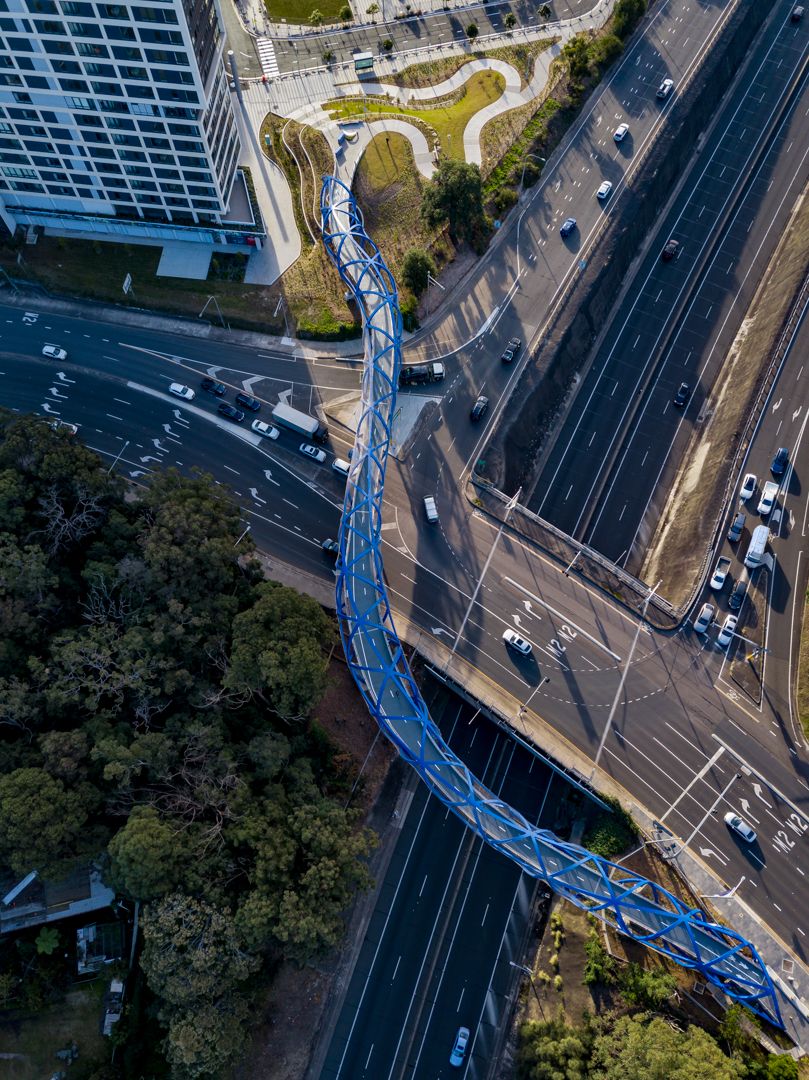 Aerial view of a curved blue steel pedestrian bridge crossing multiple busy roads next to a tall building and green trees.