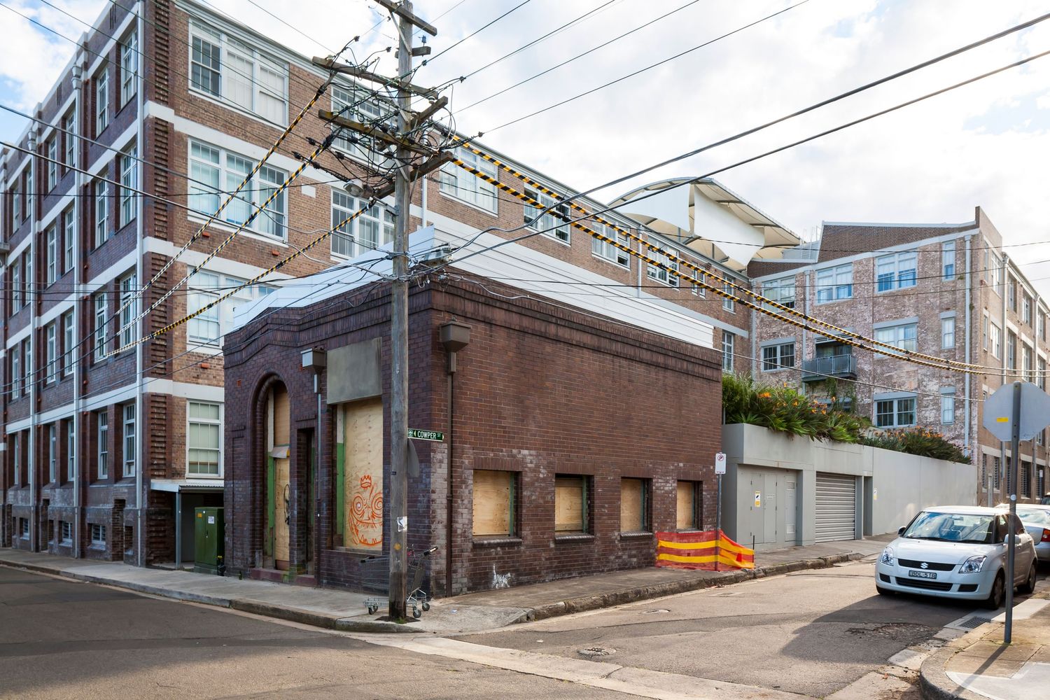 Old brick building with boarded windows on a street corner beside modern apartment blocks and overhead power lines.