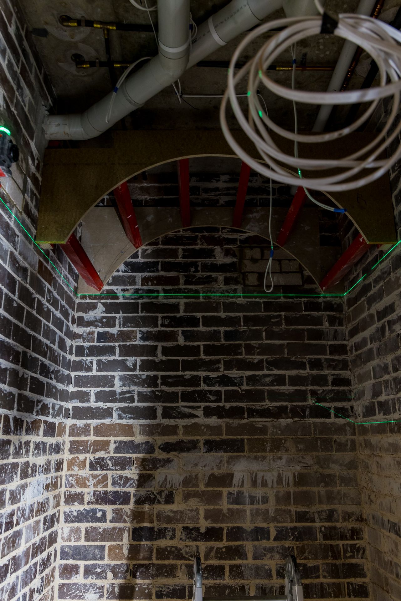 Basement brick chamber with an arched timber formwork, red steel supports, overhead pipes and coiled white cabling.