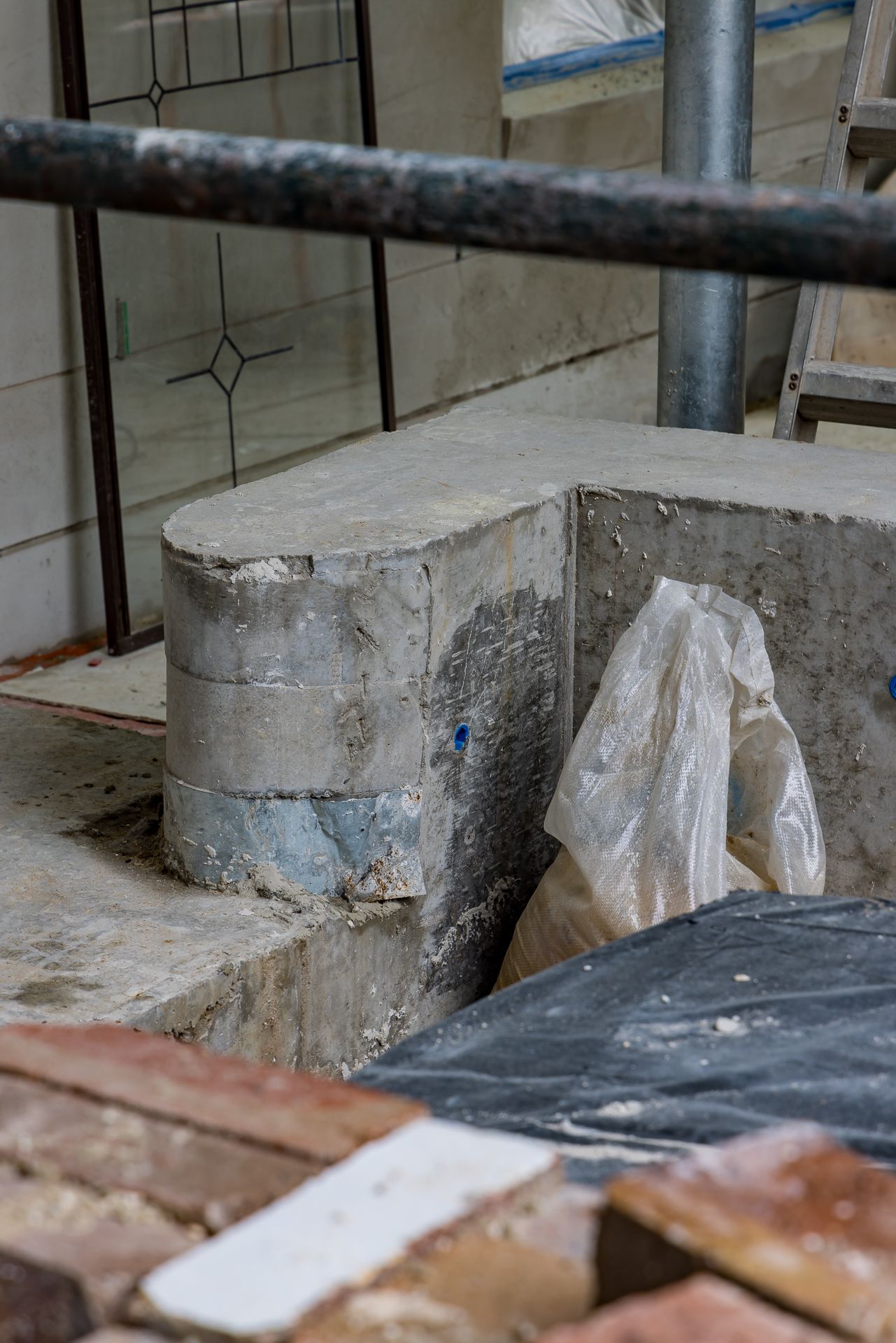 Concrete and masonry foundation detail at a renovation site with plastic sheeting, scaffolding and a leadlight window behind.