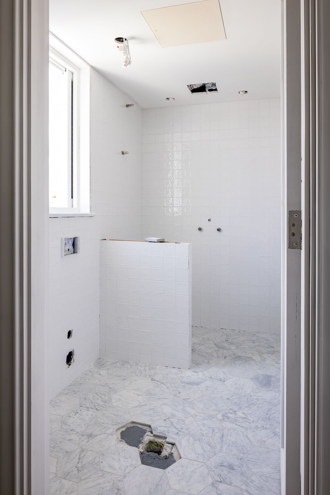 Unfinished bathroom with white square wall tiles, hexagonal marble floor tiles and exposed plumbing during renovation.