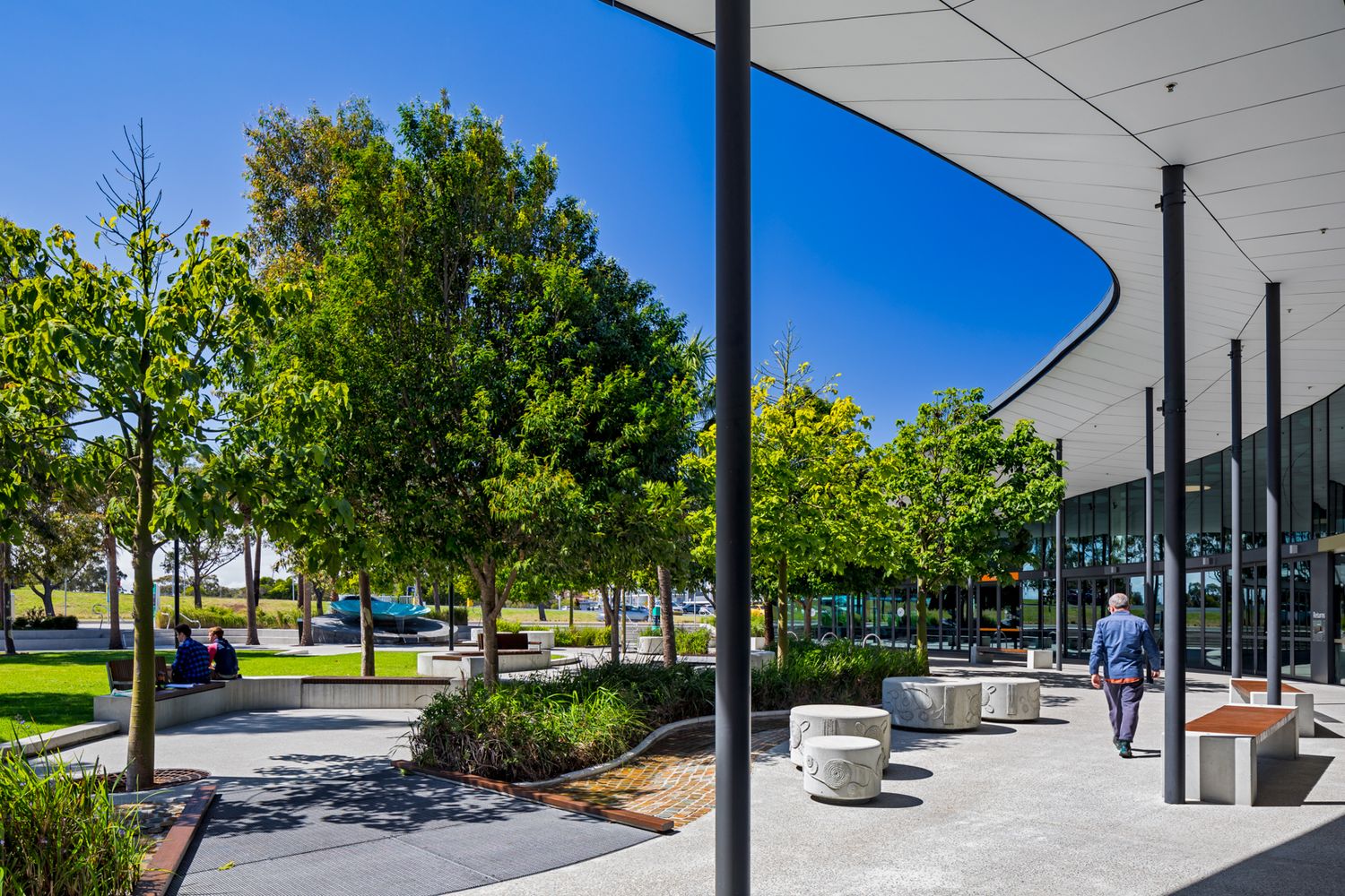 Person walks along a modern curved building canopy beside a landscaped park with mature trees, seating and green lawns.