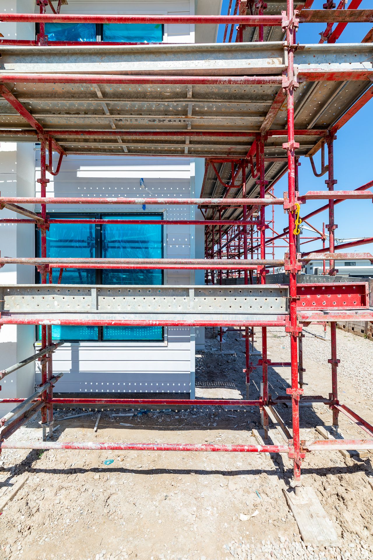 Red steel scaffolding surrounds a white building under construction with blue-wrapped windows on a gravel site.