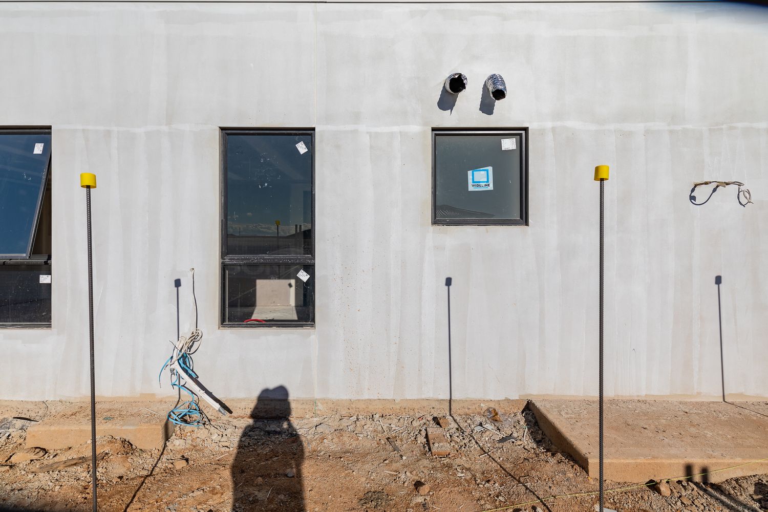 Exterior wall of a new concrete home under construction with windows, plumbing pipes, exposed cables and marker stakes in the dirt.