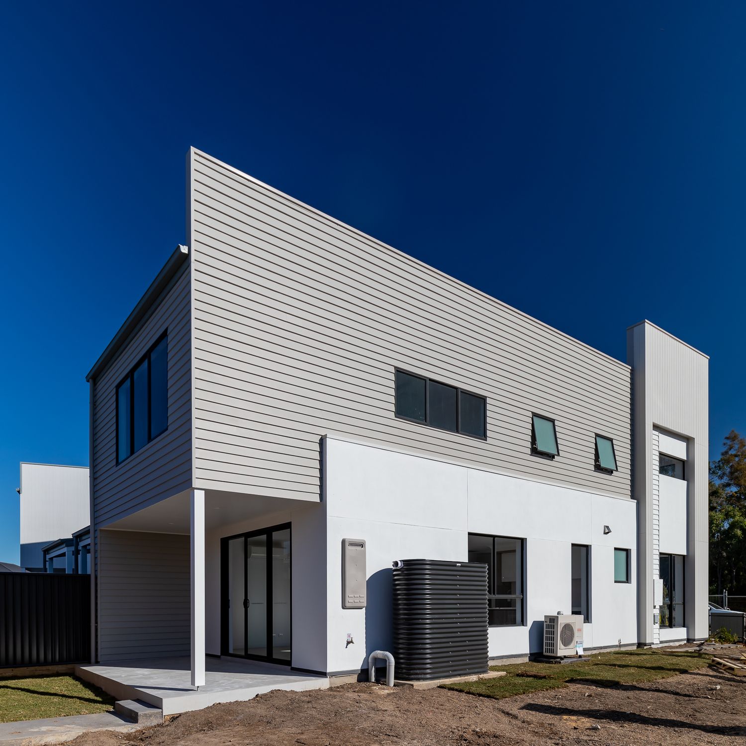 Side view of a modern two-storey home with grey cladding, white rendered walls, sliding glass doors, air conditioner and water tank.
