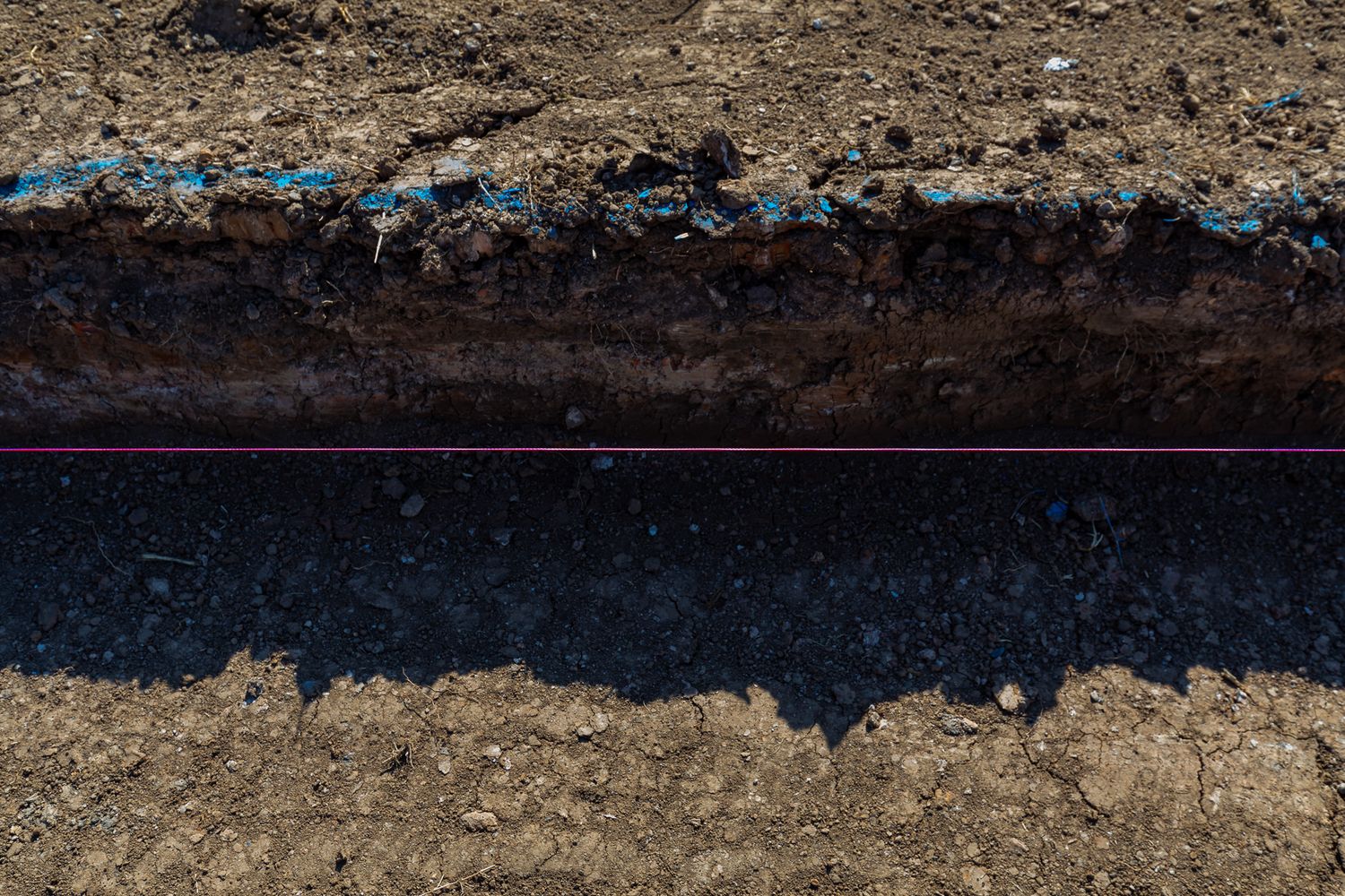 Aerial view of a freshly dug trench showing layered soil, blue pipe markings and a purple utility line in the ground.