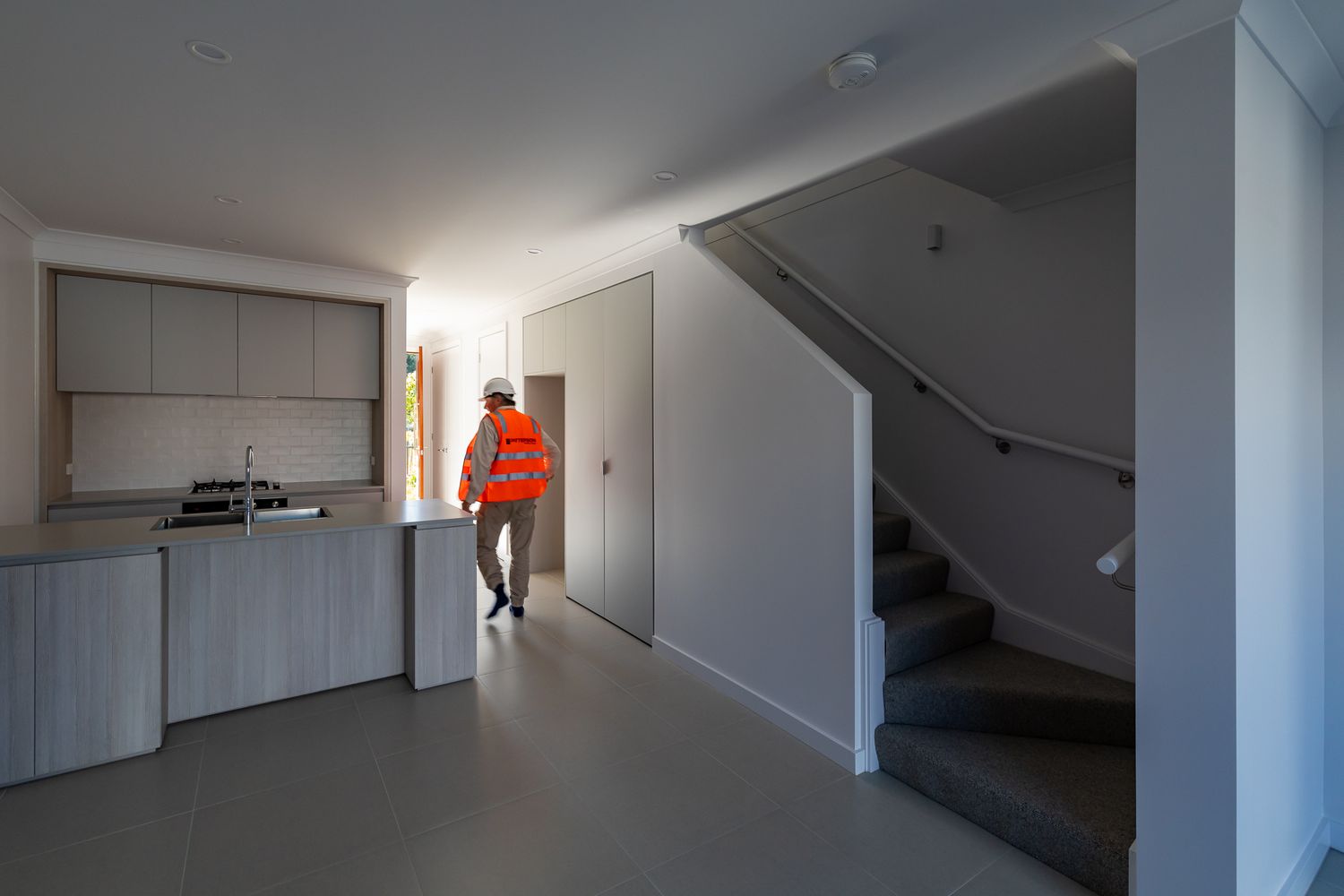 Completed townhouse interior with a kitchen bench, stairs and a worker in an orange hi-vis vest inspecting the open-plan living space.
