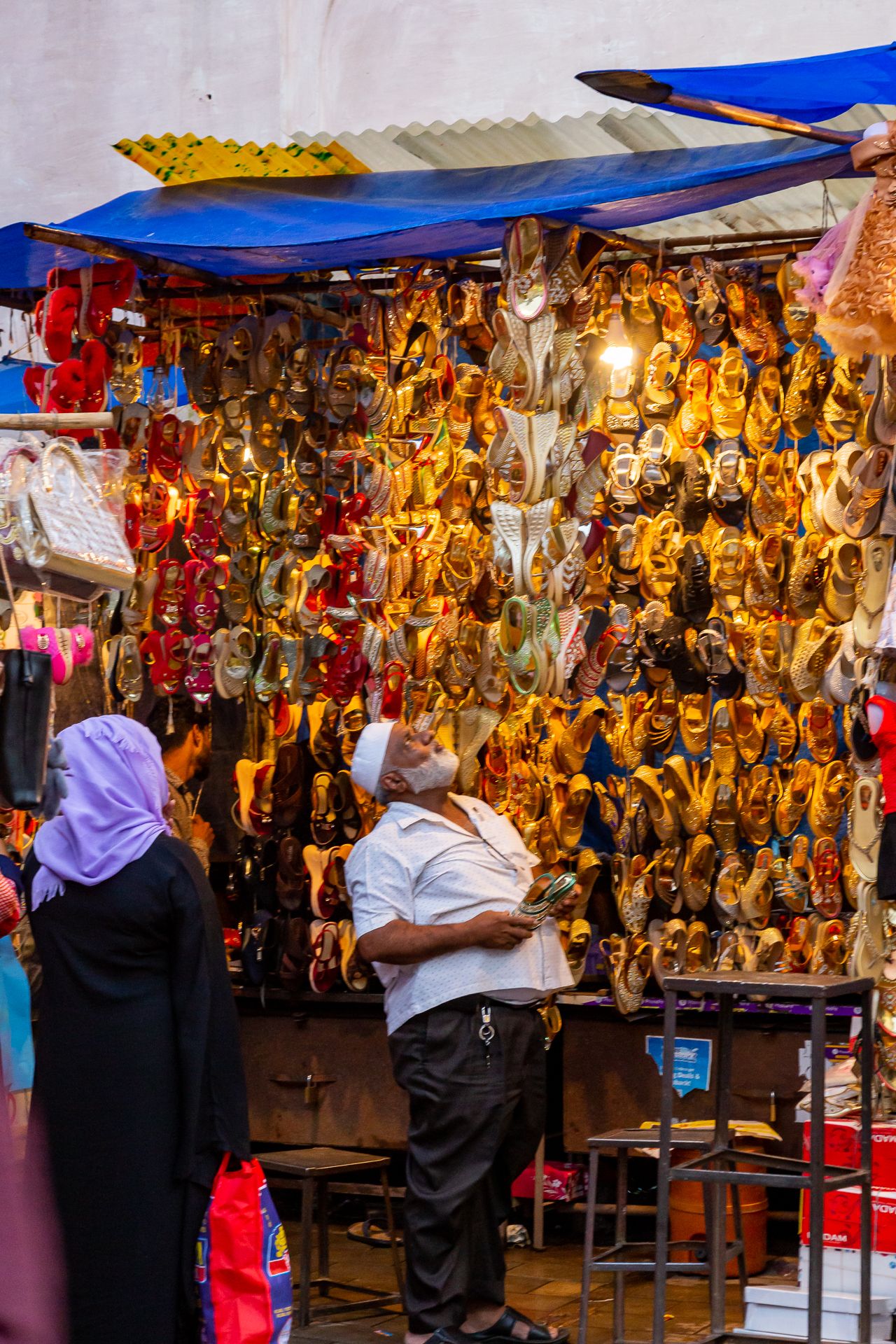 Shopper in a white cap browses a vibrant market stall packed with rows of golden and red decorative footwear under a blue tarp.