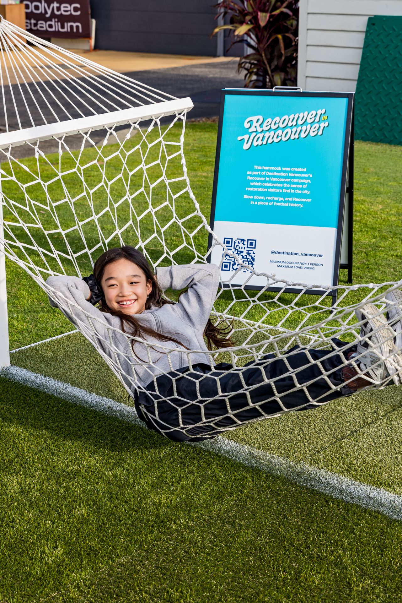Happy girl relaxes in a goalpost hammock on artificial turf next to a Recover in Vancouver information sign.