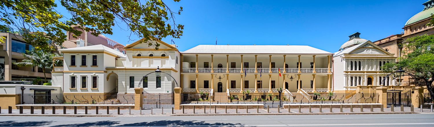 Wide view of a historic Victorian sandstone government building with a long upper verandah and ornate balustrades beneath a clear blue sky.