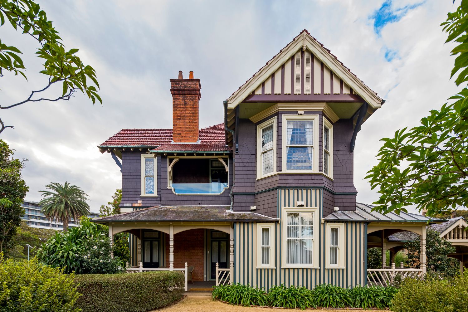 Two-story Victorian timber house with gabled roof, brick chimney, verandah, and bay windows surrounded by trees and hedges.