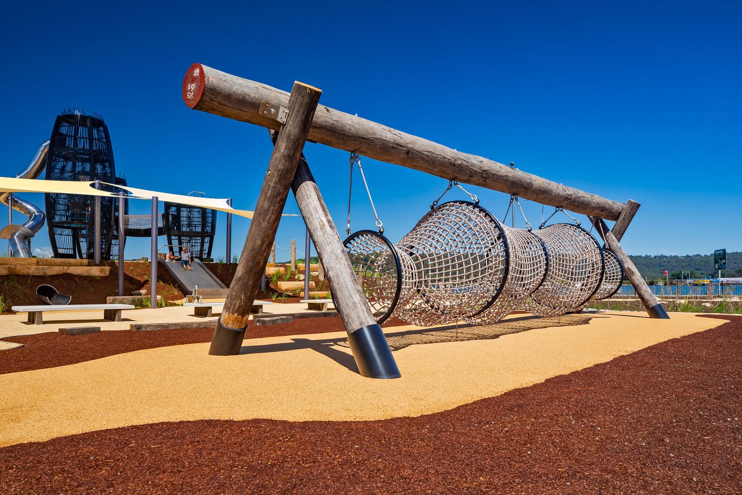 A large timber swing frame holds cylindrical rope net tunnels on a sandy playground area beside a lattice tower.