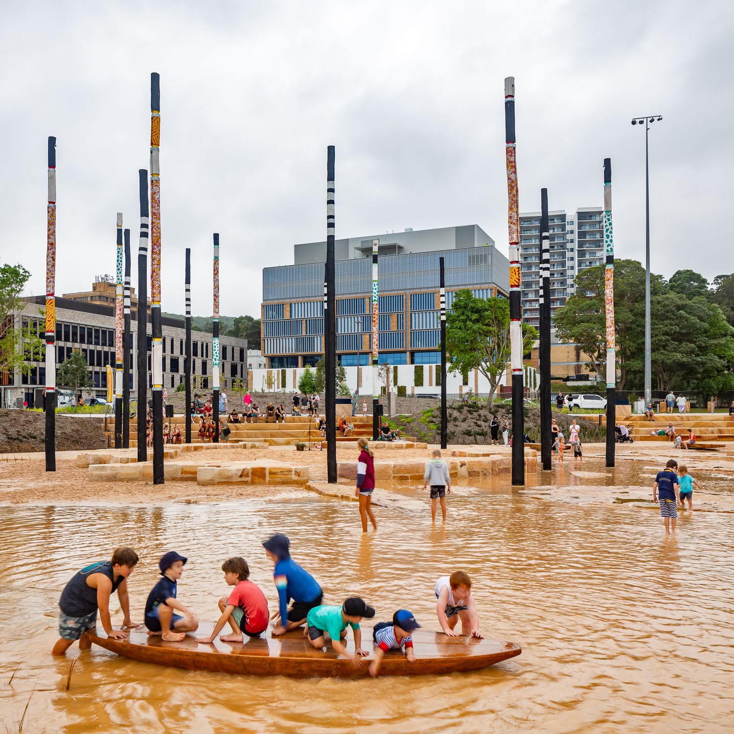 Children paddle a wooden boat in a shallow sandy water play area surrounded by tall painted poles and city buildings.