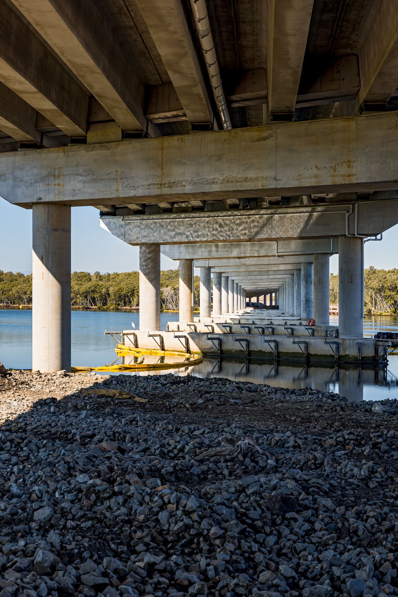 View beneath a concrete highway bridge showing rows of columns, a rocky shoreline and calm river water lined with trees.
