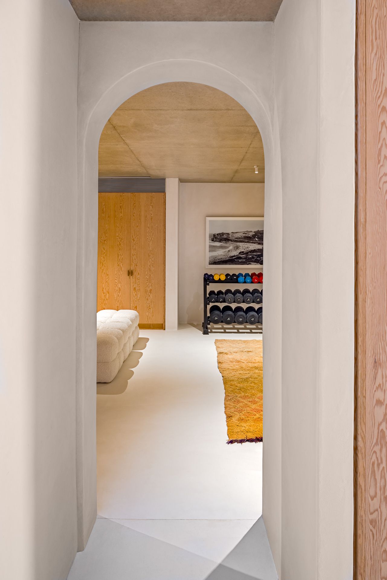 Arched doorway in a white plaster hallway leading to a home gym with a dumbbell rack, wood cabinetry and a yellow rug.