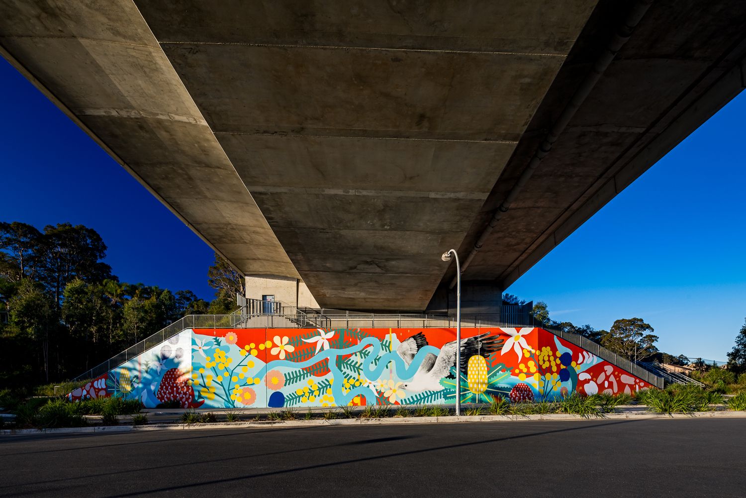 Colorful abstract mural painted on a retaining wall beneath a concrete bridge with a street lamp and trees against a blue sky.