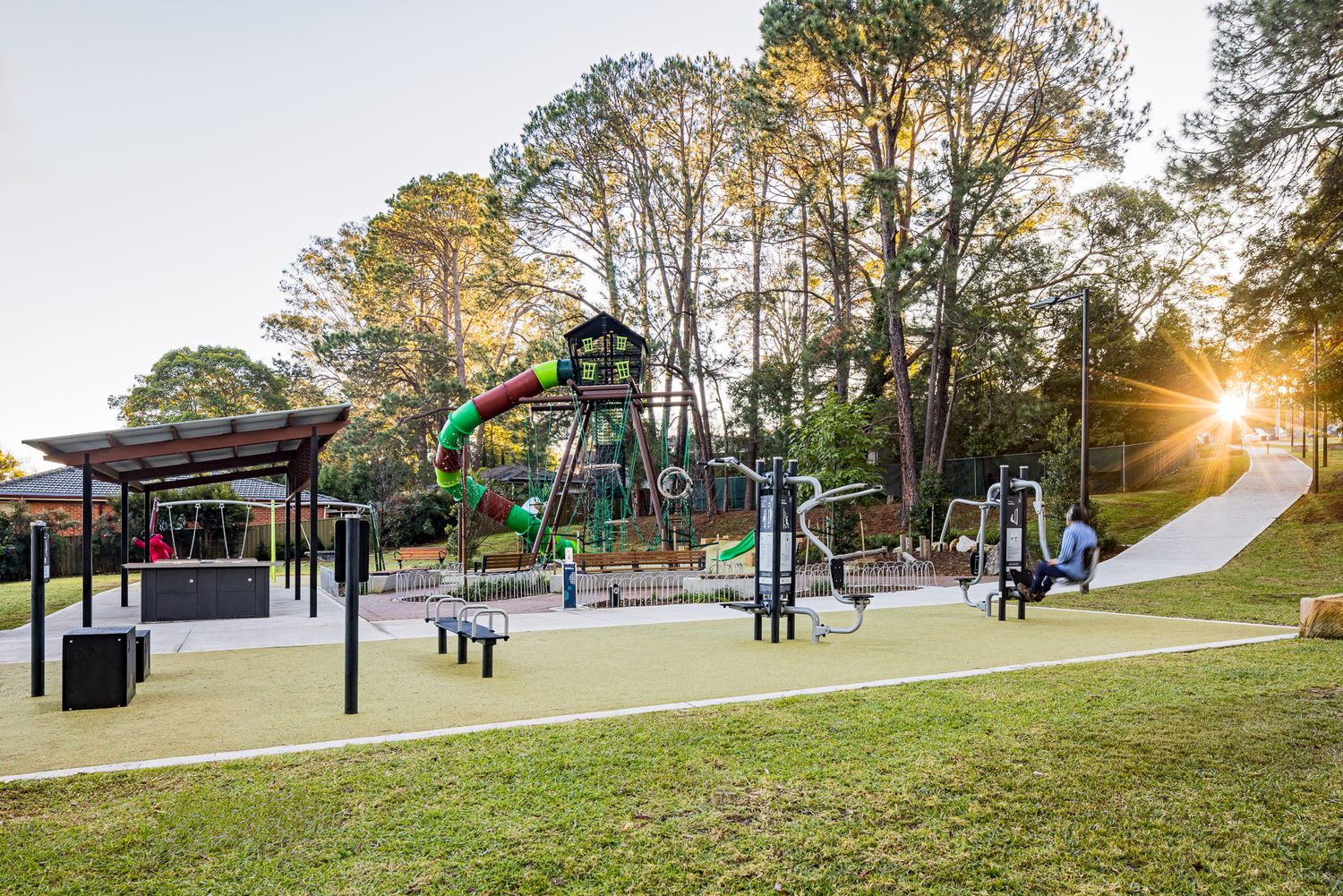 Sunset glows through tall trees over a modern playground with slides, climbing frames, fitness equipment and a shaded picnic shelter.