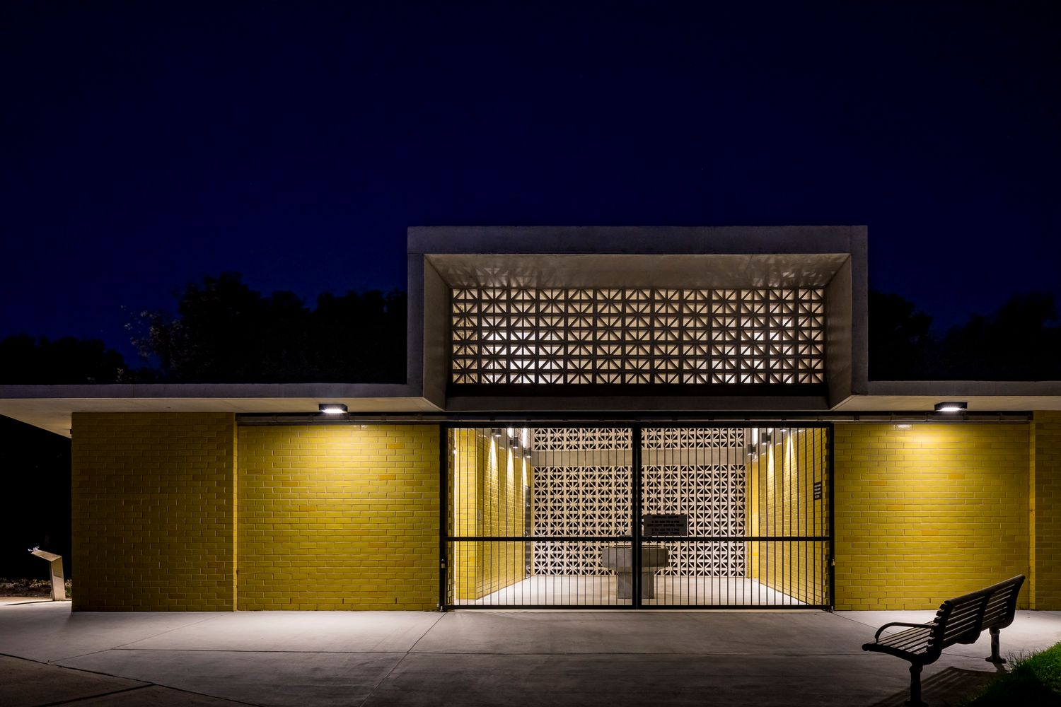 Front view of a yellow brick park amenity building at night with a patterned screen above a gated entrance and a bench nearby.