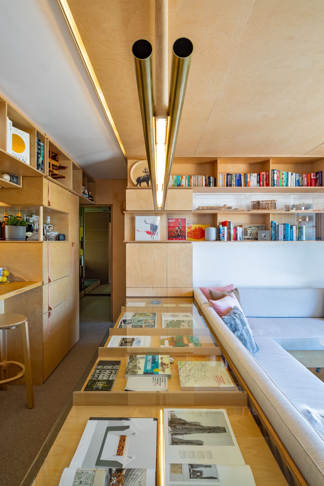 Narrow plywood apartment interior with brass pendant lights, a glass display table of books, built-in shelving, and a daybed sofa.