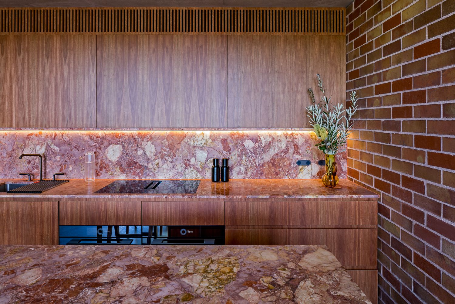 Timber kitchen cabinetry with a glowing pink marble splashback and benchtop beside a warm red brick wall.