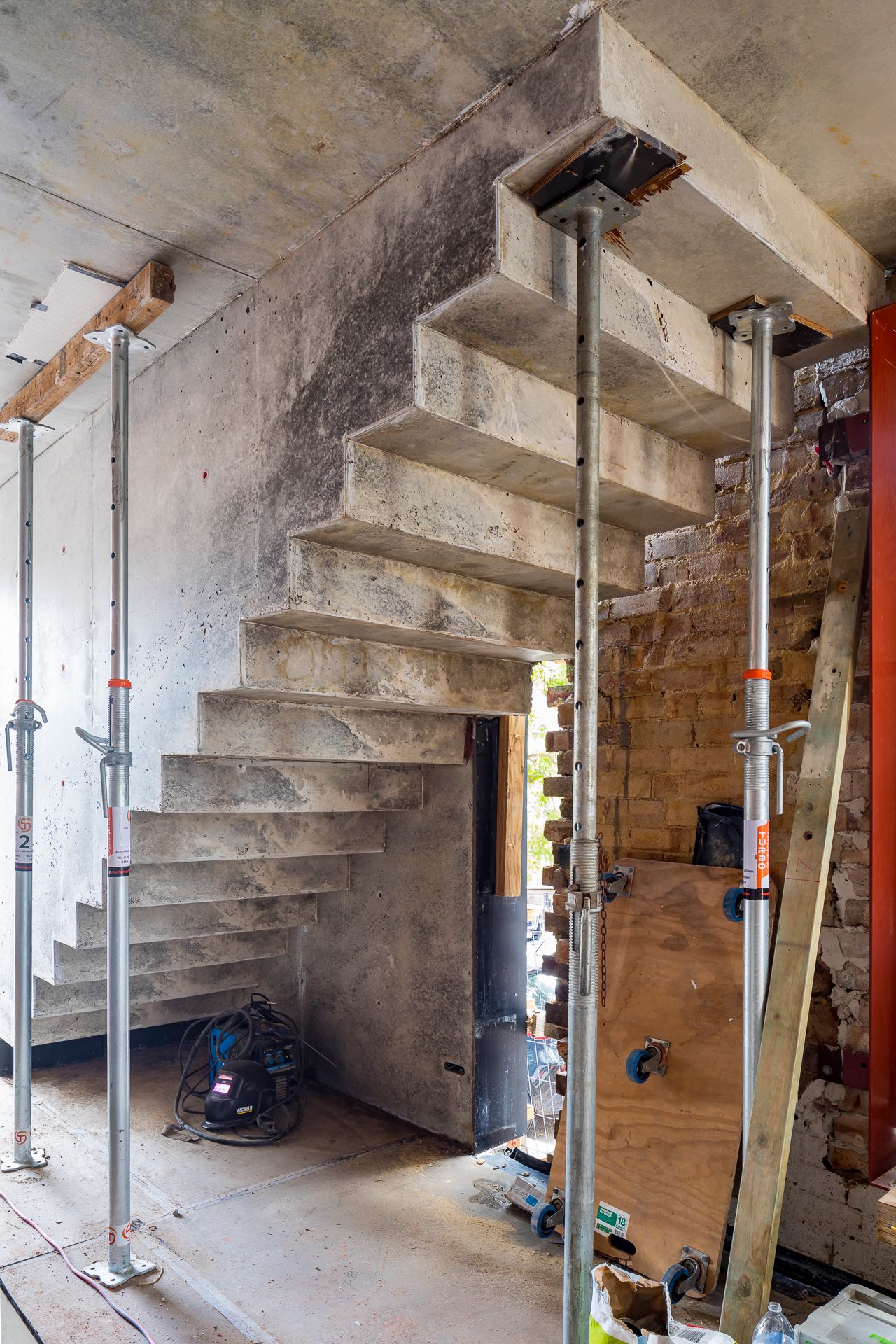 Raw concrete staircase supported by adjustable steel props inside a building under construction next to an exposed brick wall.