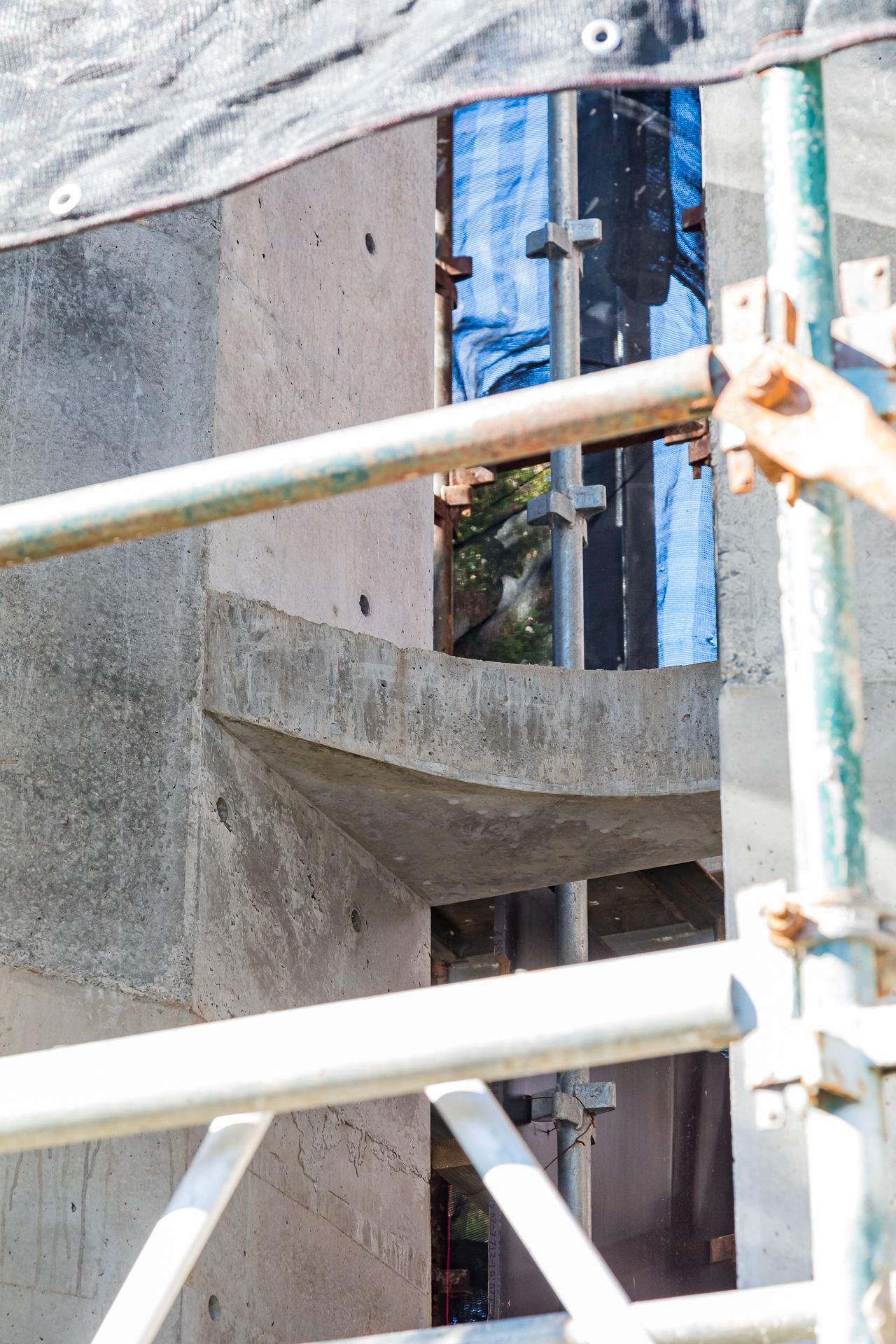 Raw concrete structure with a curved ledge seen through scaffolding poles at a building construction site.