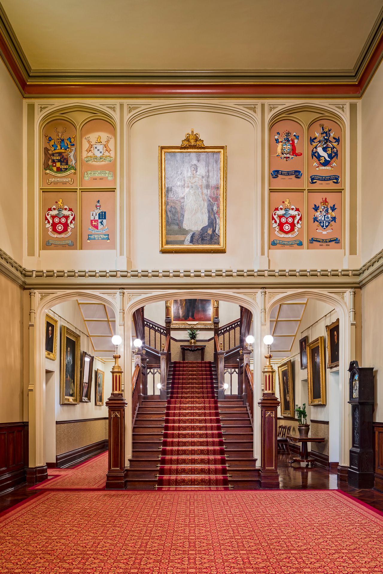 Stately hall with a red carpeted staircase, heraldic coat of arms panels, framed paintings and polished timber floors.