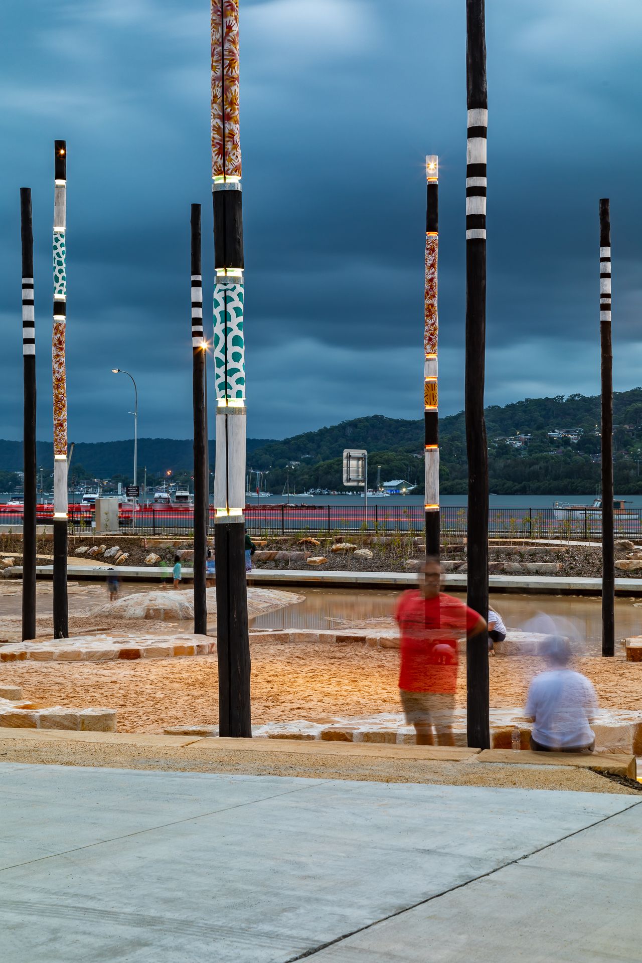 Motion-blurred visitors stand among patterned public art poles on a waterfront plaza with hills and cloudy skies behind.