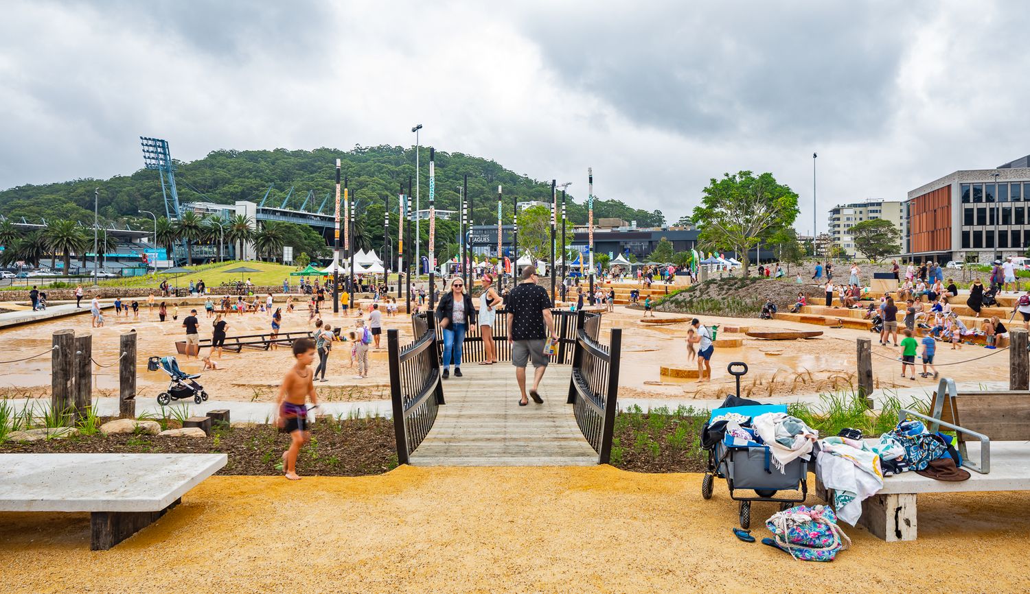 Families cross a timber bridge over native gardens toward a sandy water play area in a busy urban waterfront park.