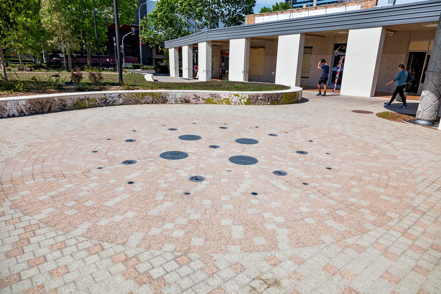 Pink brick paving with circular dark inlays forms a public courtyard beside a covered colonnade and curved stone garden wall.