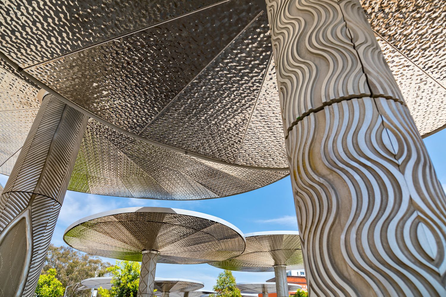 Upward view of intricately patterned metal columns and perforated canopies against a blue sky in a contemporary public space.