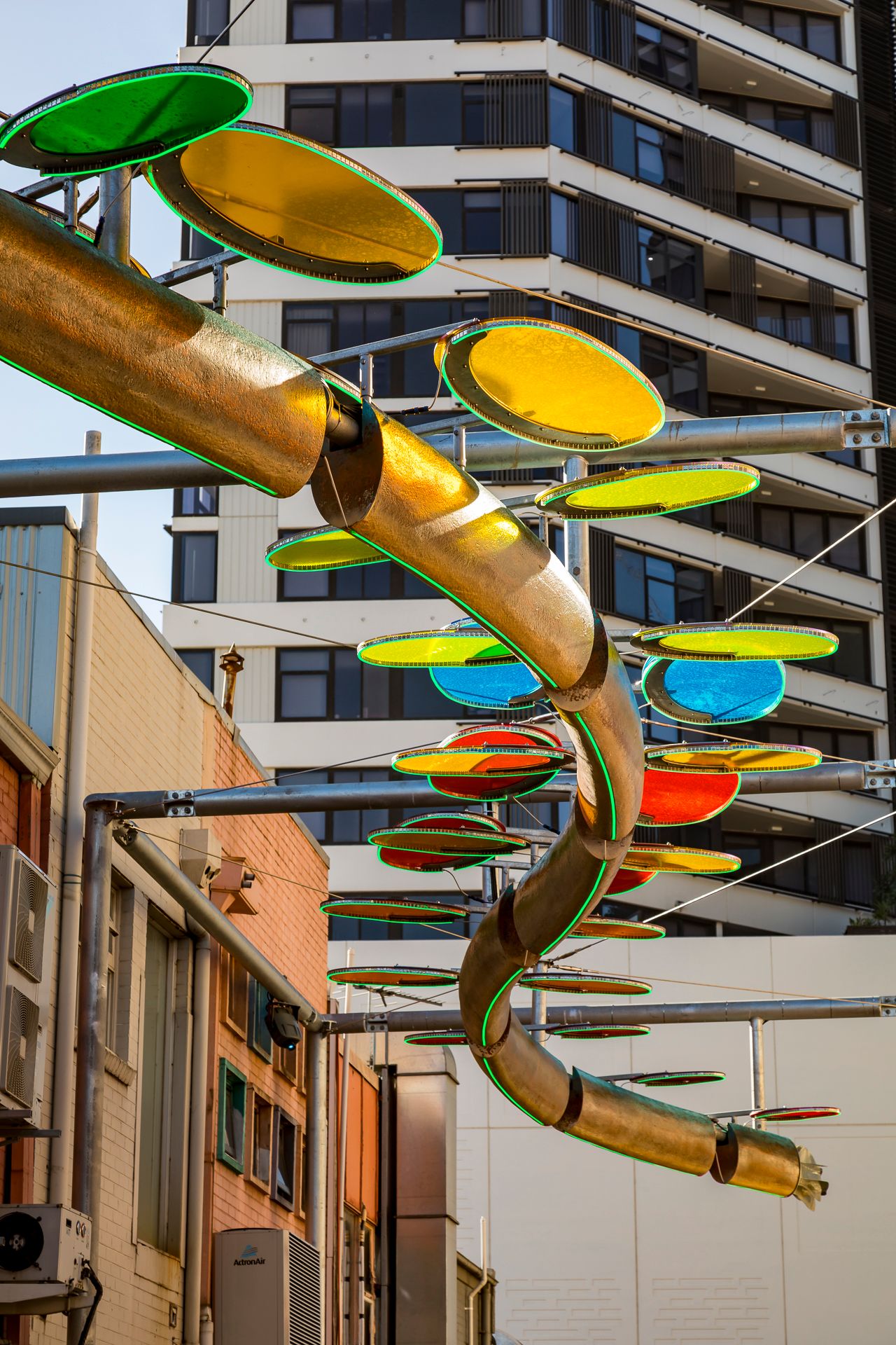 Colorful translucent disc sculpture with a curved golden tube winding between urban buildings against a blue sky.
