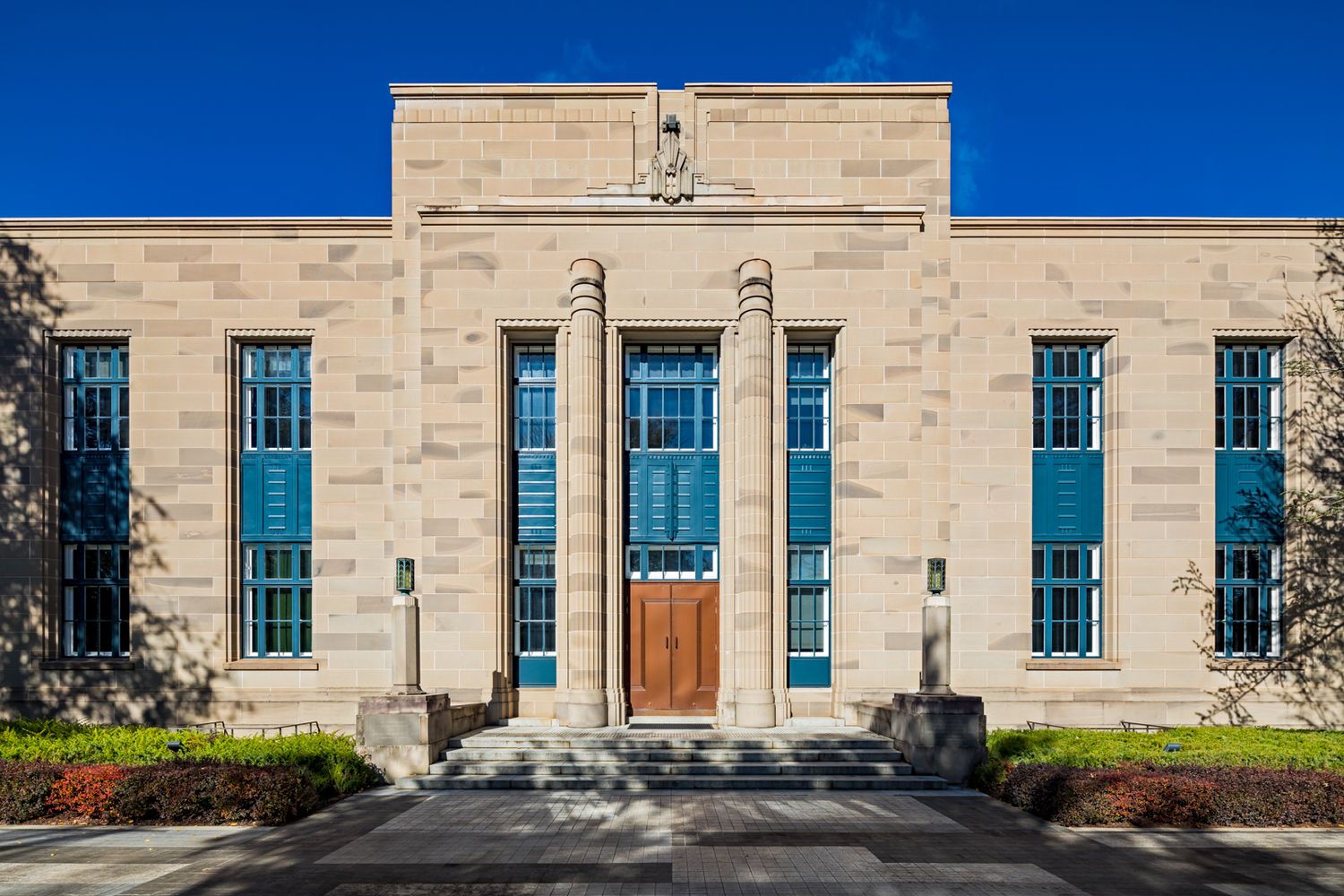 Symmetrical limestone Art Deco building facade with tall blue-tinted windows, fluted columns, and a central wooden door beneath a clear blue