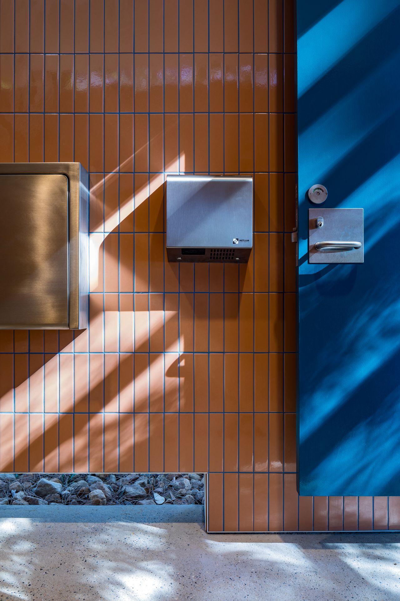 Orange tiled restroom wall with a steel hand dryer and a bright blue door, patterned by dappled sunlight shadows.