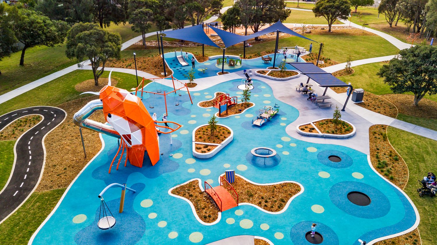 Aerial view of a colorful park playground with a blue rubber surface, orange slide, shade sails, planted islands and winding paths.