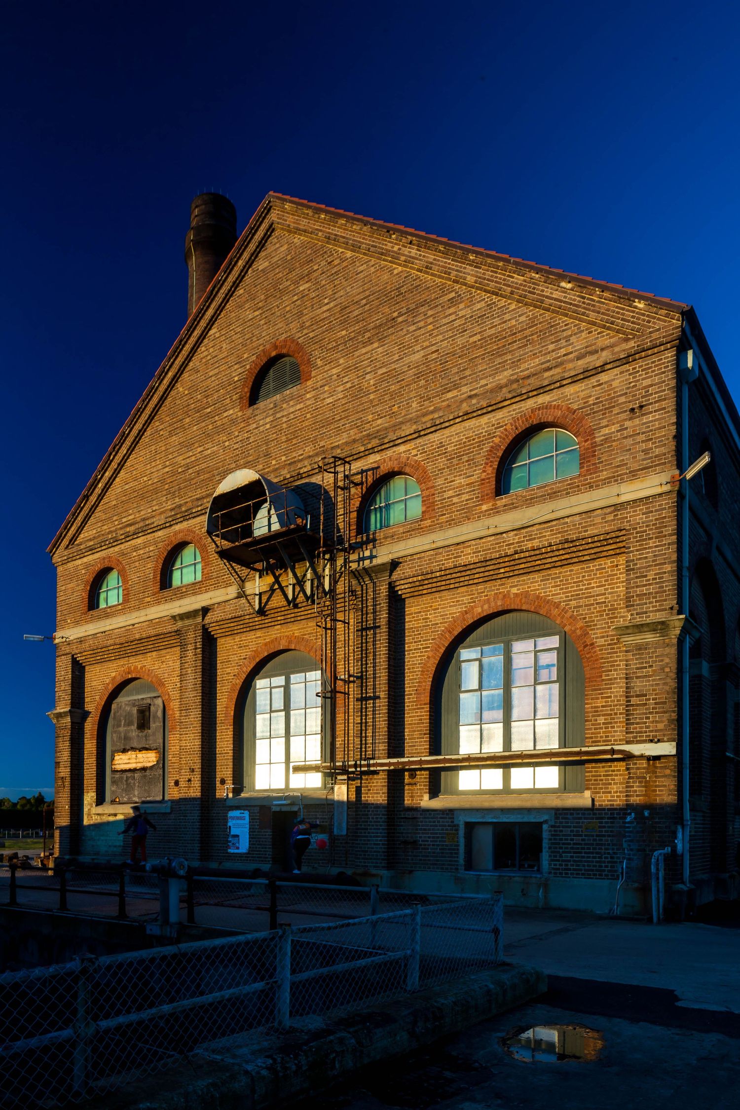 Historic red brick industrial building with arched windows and a tall chimney, glowing against a deep blue twilight sky.