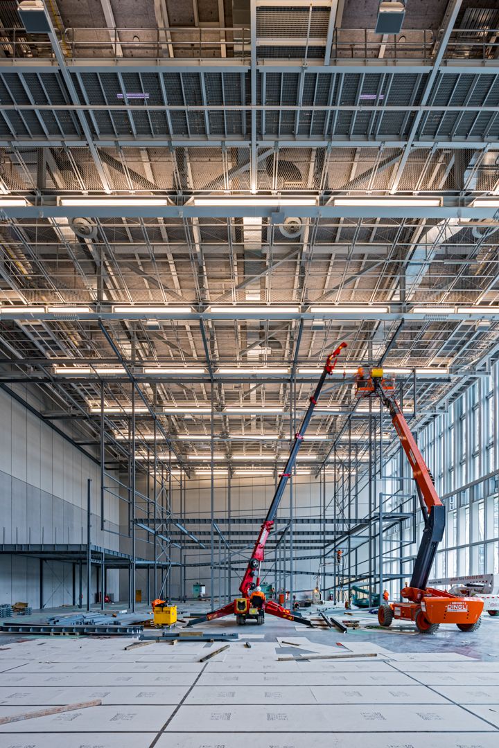 Large industrial interior under construction with steel roof trusses, scaffolding and two orange mobile lifts.