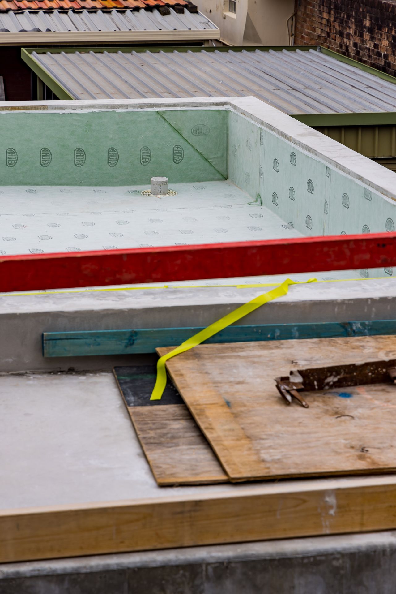 Flat roof under construction with green waterproof membrane, a red edge board, yellow tape and timber offcuts beside neighbouring rooftops.
