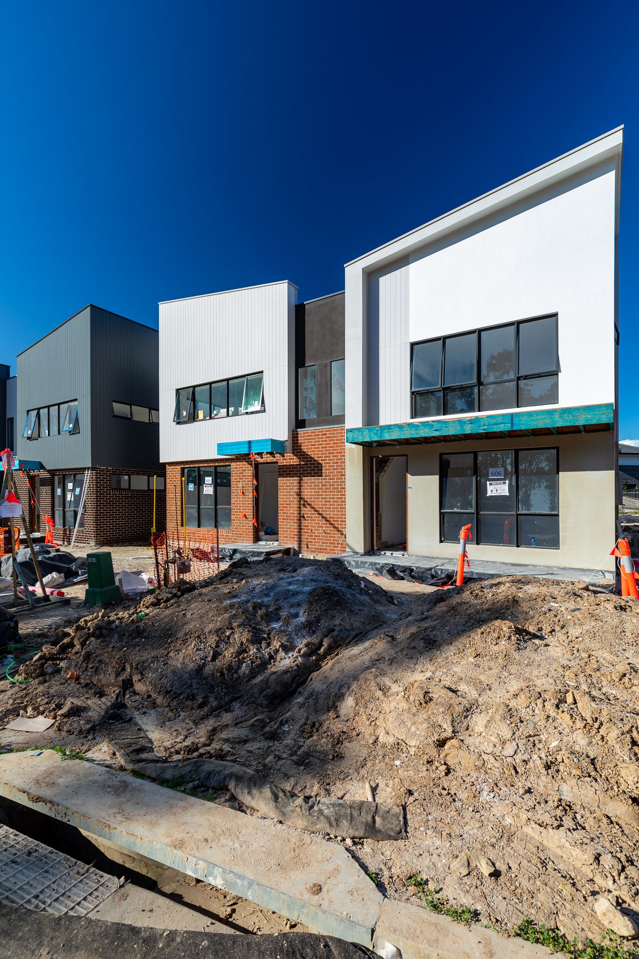 Newly built modern townhouses with white and brick facades beside a mound of excavated soil under a clear blue sky.