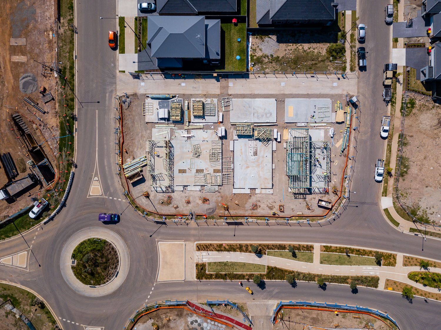 Aerial view of a residential construction site with building slabs next to a roundabout, surrounded by roads, new homes and bushland.