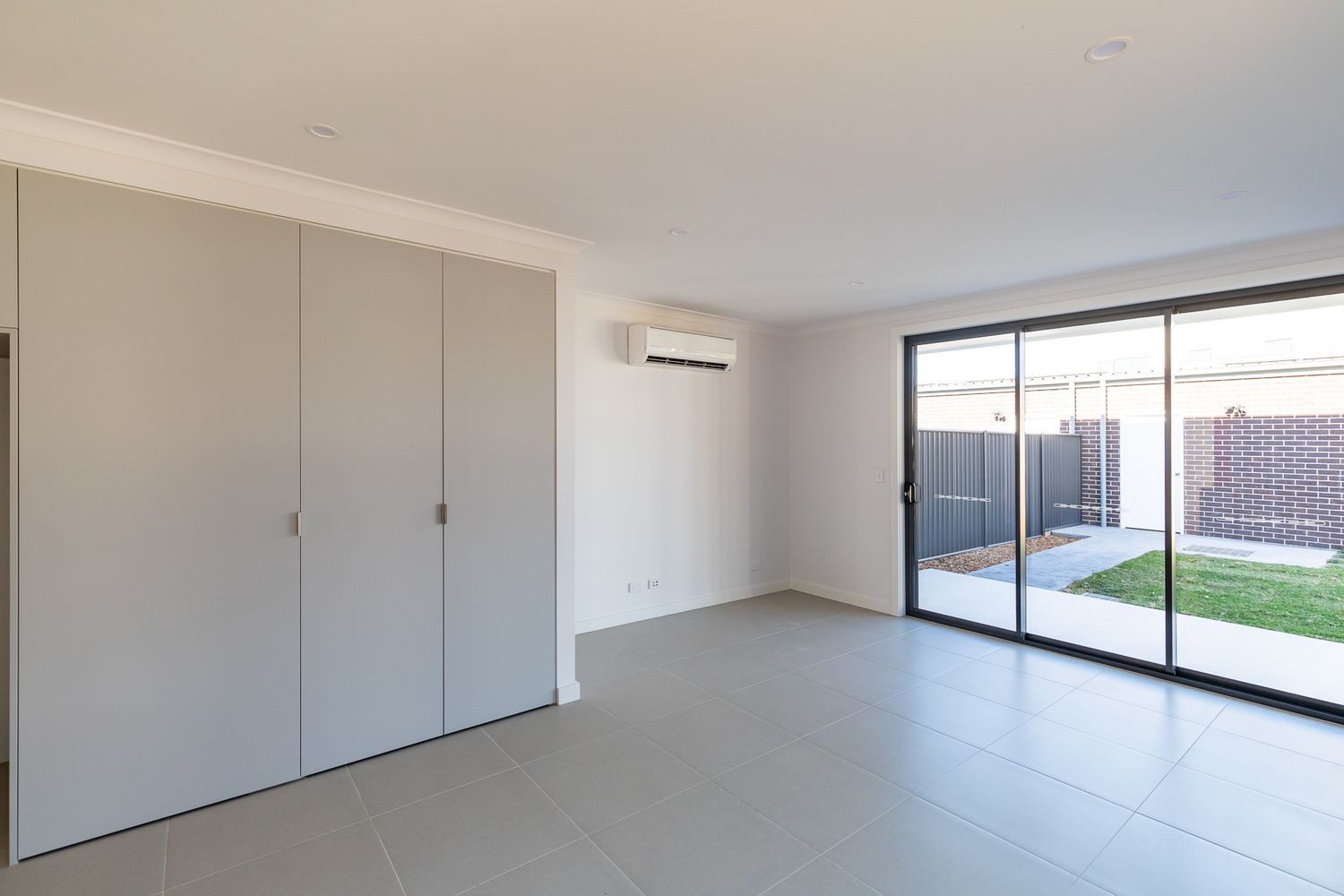 Bright empty bedroom with built-in wardrobes, a split-system air conditioner and sliding glass doors opening to a fenced backyard.